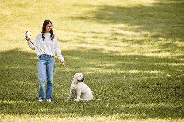 Samimi sonbahar kıyafetleri içinde bir kadın neşeli köpeğiyle yemyeşil bir parkta geziniyor..