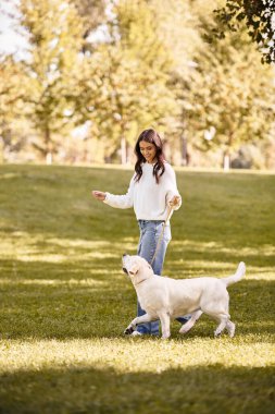 A young woman in autumn attire joyfully walks her dog through a lively park filled with trees. clipart