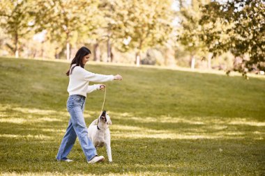 Sıcak bir sonbahar kıyafetine sarılmış genç bir kadın, köpeğiyle parkta neşeyle geziniyor..