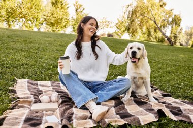 Genç bir kadın battaniyeye oturmuş, mutlu bir köpeğin yanında kahvesinin tadını çıkarırken gülümsüyor..