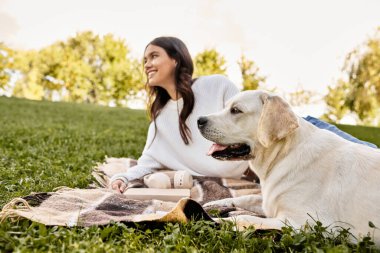 Genç bir kadın güneşli bir parkta battaniyeye oturmuş köpeğiyle rahatlarken gülümsüyor..
