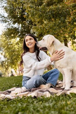 Samimi giyinmiş genç bir kadın güneşli bir parkta köpeğiyle neşeyle etkileşime girer..