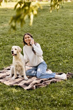 Neşeli genç bir kadın, köpeği ile parkta battaniyenin üzerinde otururken telefonuyla sohbet ediyor..