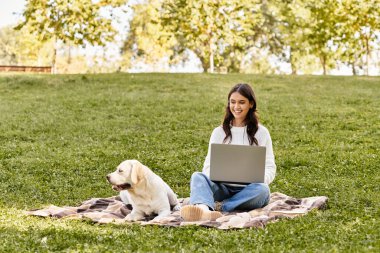 Sonbahar için giyinmiş genç bir kadın, dizüstü bilgisayarı ve sadık köpeğiyle parkta dinleniyor..