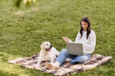 Sıcak sonbahar kıyafetleri içinde neşeli genç bir kadın köpeğiyle bir battaniyeye uzanıyor ve dışarıda çalışıyor..