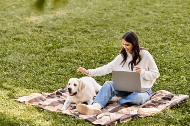 Genç kadın parkta bir battaniyenin üzerinde oturuyor, köpeğiyle oynaşırken dizüstü bilgisayarında yazıyor..