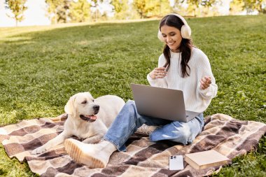 Sıcak giysili genç bir kadın battaniyenin üzerinde oturuyor. Yanında köpeğiyle laptopunu neşeyle kullanıyor..