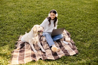 Huzurlu bir parkta, sıcak giyimli genç bir kadın dizüstü bilgisayarıyla çalışıyor, köpeğiyle neşe paylaşıyor..