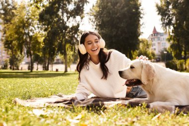 Neşeli genç bir kadın çimenlerin üzerinde rahatlar, canlı bir parkta köpeğiyle mutlu bir şekilde etkileşim içindedir..