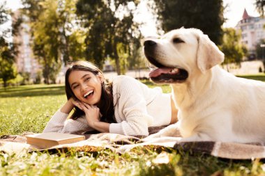 Güzel bir kadın parkta bir battaniyeye uzanmış, köpeği yanında yatarken gülümsüyor..