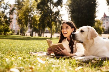 Samimi sonbahar kıyafetleri içinde genç bir kadın çimenlerin üzerinde oturuyor, yanında köpeğiyle mutlu bir şekilde kitap okuyor..