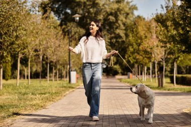 Sıcak kıyafetler giyen genç bir kadın, sonbaharda güneşli bir parkta köpeğiyle neşeyle gezinir..