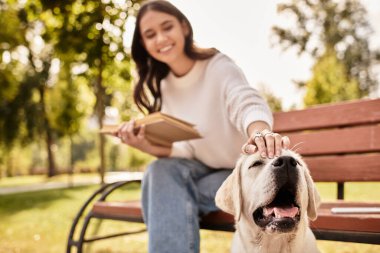 Canlı bir parkta, rahat giyimli genç bir kadın doğaya dalmış gülümseyen köpeğini sevgiyle besliyor..
