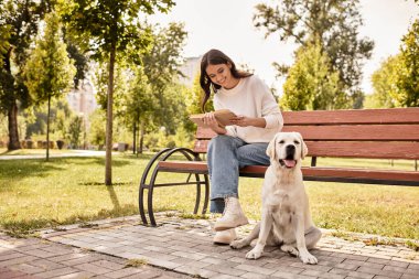 Güneşli bir parkta, genç bir kadın bir bankta oturuyor, köpeğiyle vakit geçiriyor..