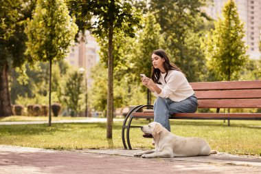 Bir bankta otururken, rahat bir sonbahar kıyafeti giymiş bir kadın köpeği yakınlarda dinlenirken telefonuna bakar..