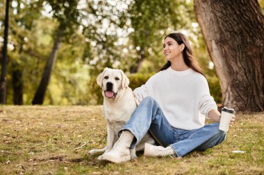 Samimi bir sonbahar elbisesi giymiş genç bir kadın neşeli köpeğiyle çimenlerde rahatlar ve kahvesini yudumlar..