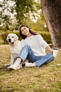 Samimi bir sonbahar kıyafeti giymiş genç bir kadın köpeğinin yanındaki çimenlerde oturmuş kahvesini yudumluyor..