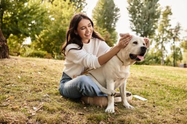 Samimi sonbahar kıyafetleri içinde mutlu bir kadın köpeğiyle altın ağaçların altında oynuyor..