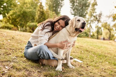 Canlı bir park ortamında, sonbahar kıyafetli genç bir kadın köpeğiyle neşeli bir anı paylaşır..