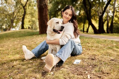 Genç bir kadın, sakin bir sonbahar parkında çimlerin üzerinde otururken sadık köpeğini kucaklıyor..