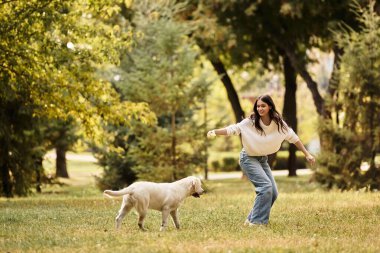Hoş bir sonbahar elbisesi içinde neşeli genç bir kadın canlı bir parkta köpeğiyle oynuyor..