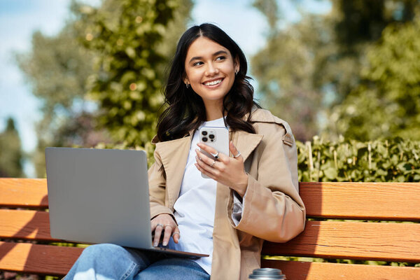 A cheerful young woman sits on a bench in the park, engaged with her laptop and smartphone.