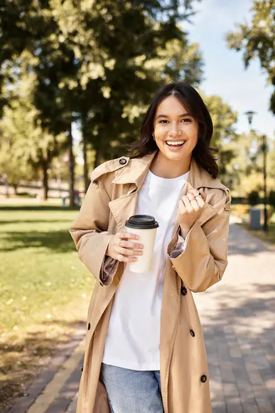 stock image A young woman in cozy autumn attire radiates happiness while holding a warm drink in the park.