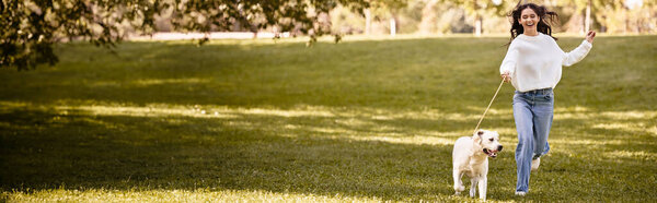A young woman in cozy autumn attire happily strolls with her dog through a sunlit park.
