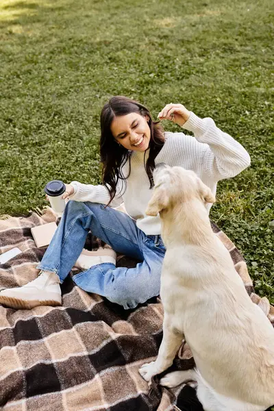 Sıcak giyimli genç bir kadın çimenli bir tarlada köpeğiyle neşeyle etkileşime girer..