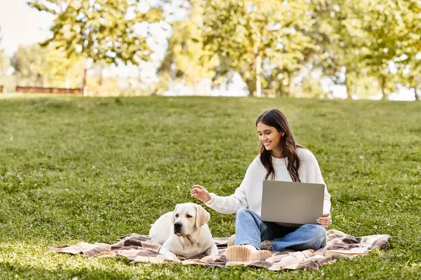 Genç bir kadın parkta bir battaniyeye oturmuş, köpeği yanında dinlenirken dizüstü bilgisayarıyla meşgul olmuş..