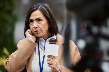 A multiracial female journalist in a beige coat focuses intently as she holds a microphone during a news report. clipart