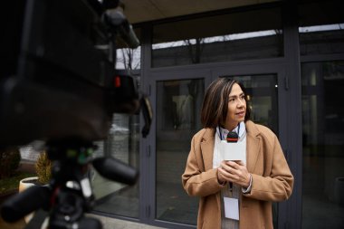 Elinde mikrofon tutan bej ceketli bir gazeteci zarif bir binanın dışında haber yapmaya hazırlanıyor..