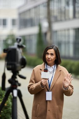 Havalı bej ceketli çok ırklı bir gazeteci açık hava mekanından tutkuyla bildiriyor..