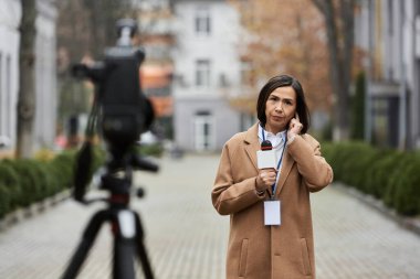 Bej ceketli çok ırklı bir kadın gazeteci sonbahar şehir manzarasında haber sunmaya hazırlanıyor..