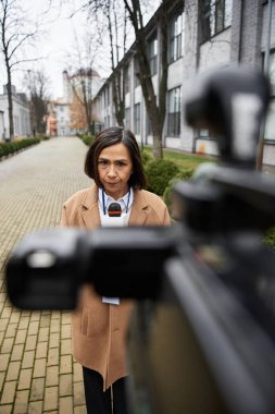 Bej ceketli, çok ırklı bir kadın gazeteci elinde mikrofonla güvenle haberleri paylaşmaya hazır bekliyor..