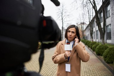 Çok ırklı bir kadın gazeteci, serin bir günde hava durumunu sunmaya hazırlanırken elinde bir mikrofon tutuyor..