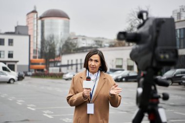 Bej ceketli çok ırklı bir gazeteci otoparkta duruyor ve kendinden emin bir şekilde bir haber sunuyor..