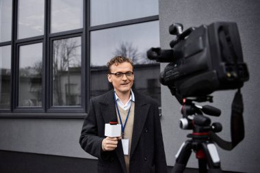 A professional journalist in glasses speaks into a microphone while standing near a camera outdoors. clipart