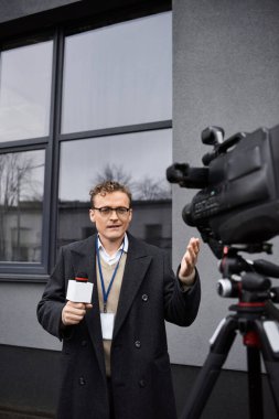 A news anchor in a coat and glasses passionately speaks into a microphone while outside a building. clipart