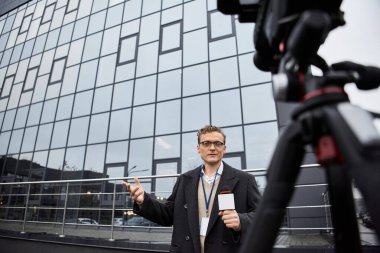 Odaklanmış bir gazeteci, elinde mikrofon tutarken cam bir binanın dışında yaşanan olayları tartışıyor.