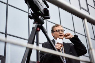 A journalist wearing glasses and a coat is speaking into a microphone, engaged in a news report outside. clipart