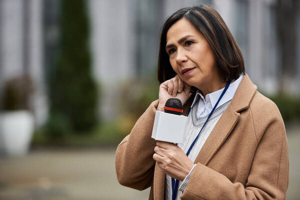 Многорасовая журналистка в бежевом пальто держит микрофон и пишет репортажи на открытом воздухе..