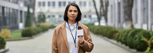 A multiracial woman in a beige coat interviews outdoors, holding a microphone in a city courtyard.