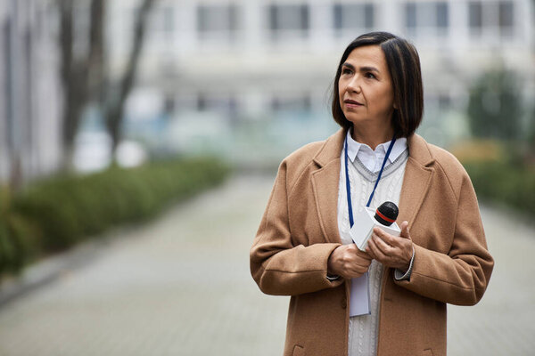 The journalist in a beige coat stands poised with a microphone, ready to share breaking news in a bustling area.