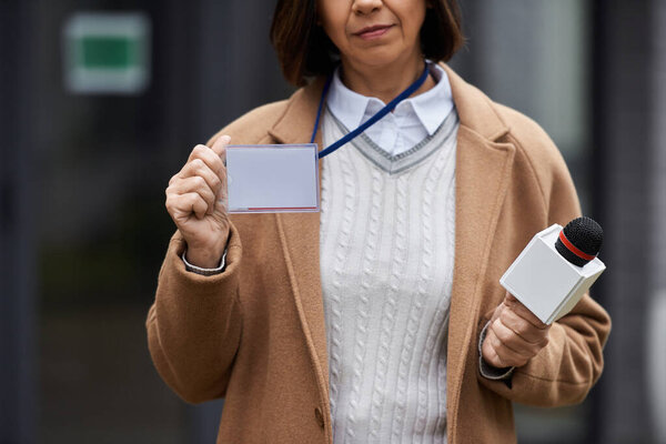 A multiracial female journalist holds a microphone while preparing to deliver a news report in the city, displaying her press badge