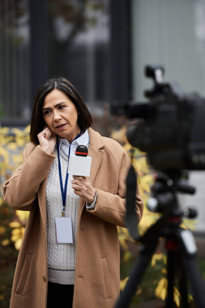 A focused multiracial female journalist holds a microphone while preparing for an outdoor news segment.