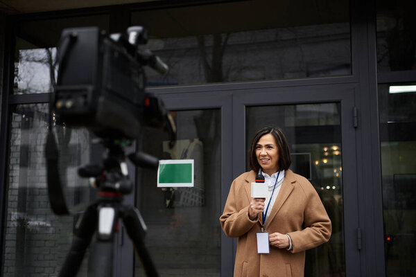 A multiracial female journalist in a beige coat actively shares news updates while holding a microphone outdoors.