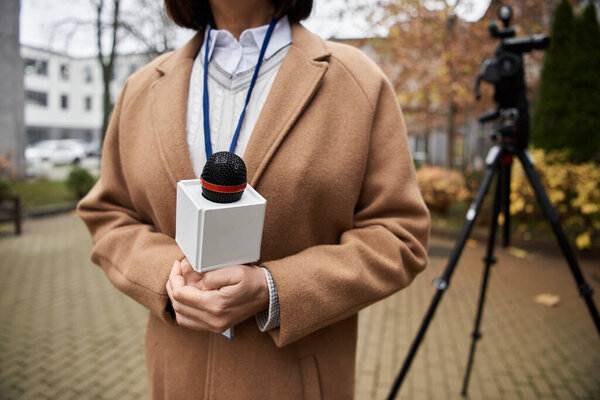 A focused journalist holding a microphone stands outdoor, preparing to deliver a news report amid autumn foliage.
