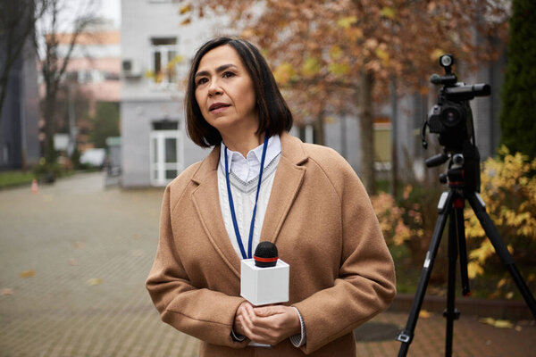 Multiracial journalist in a beige coat engages the audience with a microphone in a calming autumn atmosphere.