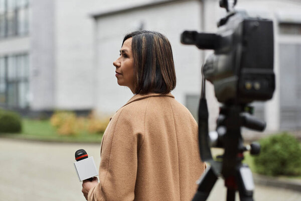 A multiracial journalist in a beige coat holds a microphone, preparing to report on current events outdoors.